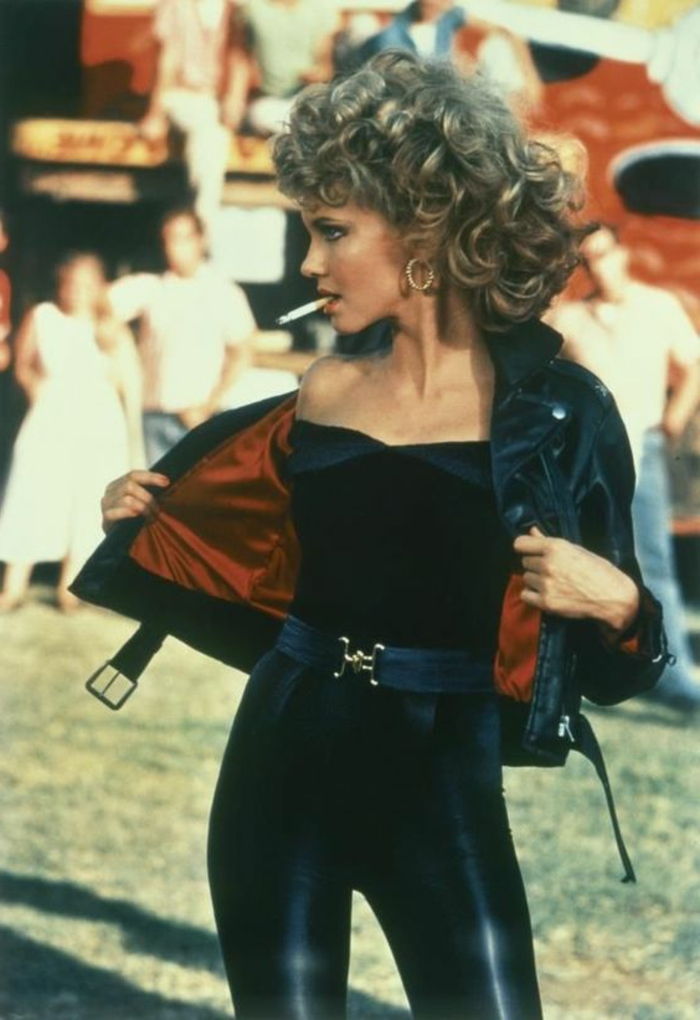 Roupa dos anos 80 de Olivia Newton John com calça de couro preta, blusa sem alças preta e jaqueta de couro preta