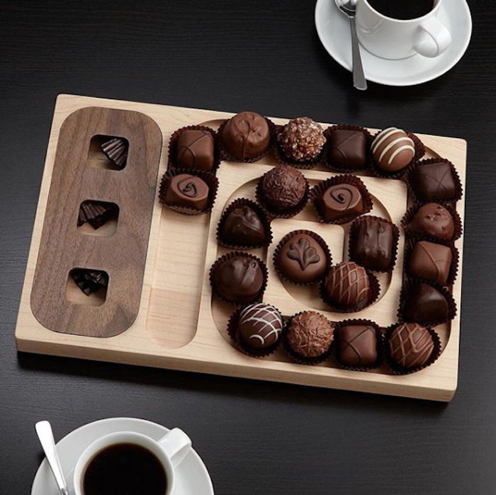 Drewniany półmisek z eleganckim wzorem, czekoladki czekoladowe z mleczną i ciemną czekoladą, trzy małe kwadratowe otwory, dwie porcelanowe filiżanki z czarną kawą, dwie łyżeczki na czarnym stole
