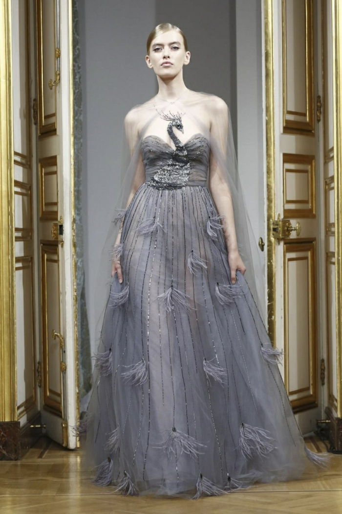 Yanina Couture Collection - elegantna dolga siva brez oblačil iz lahkih tkanin z aplikacijami, nagubana na vrhu z motivom pave, oblečena s prozornim tulle šal