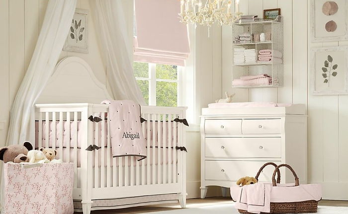 Kūdikių kambarys su švelniu elegantišku, baltos medienos baldais, šviesiai rausva patalynė, žaisminga liustra