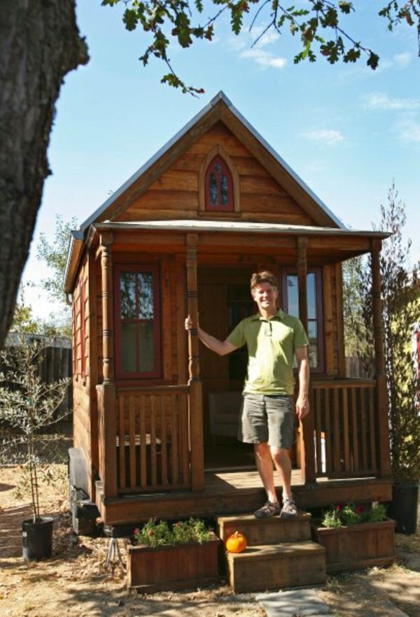 casă mică din lemn cu acoperiș interesant și un bărbat în fața lui