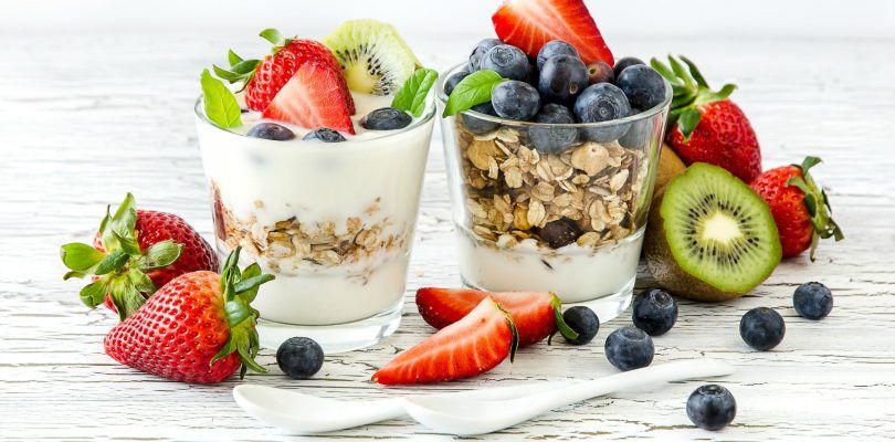 Yoghurt i glas med tre sorter av frukt - kiwi, jordgubbar, blåbär, serveras med havregryn, vitmålade träbord, två små porslinssked