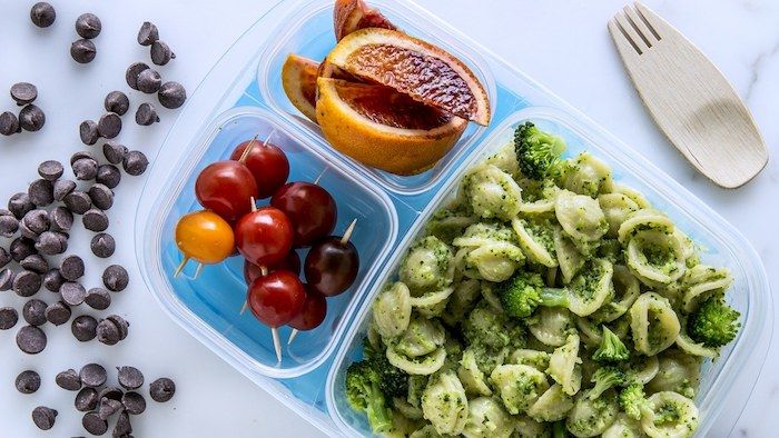 Pasta lunchbox med grön pesto och broccoli, tre typer av körsbärstomater, stekt apelsiner, chokladdroppar, trägaffel, träsked