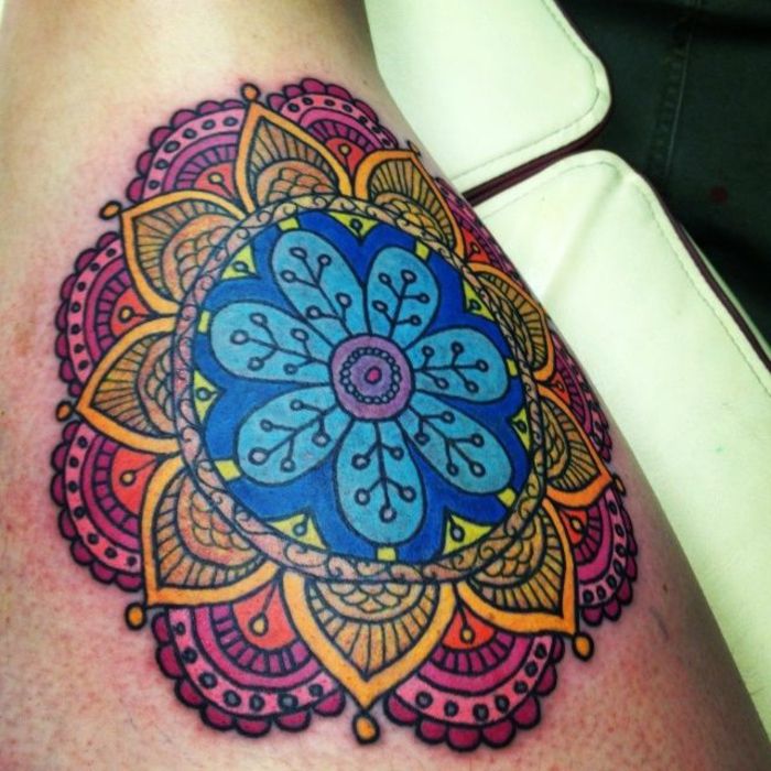 tatuagem de mandala colorida em quatro cores - fora violeta e laranja, dentro de amarelo e azul, motivos florais em muitas cores, espreguiçadeira de couro branco