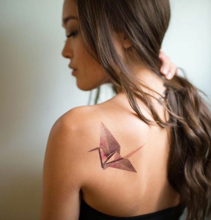 Ecco una giovane donna con un tatuaggio origami sulla sua scapola - un tatuaggio origami con un uccello origami volante rosso