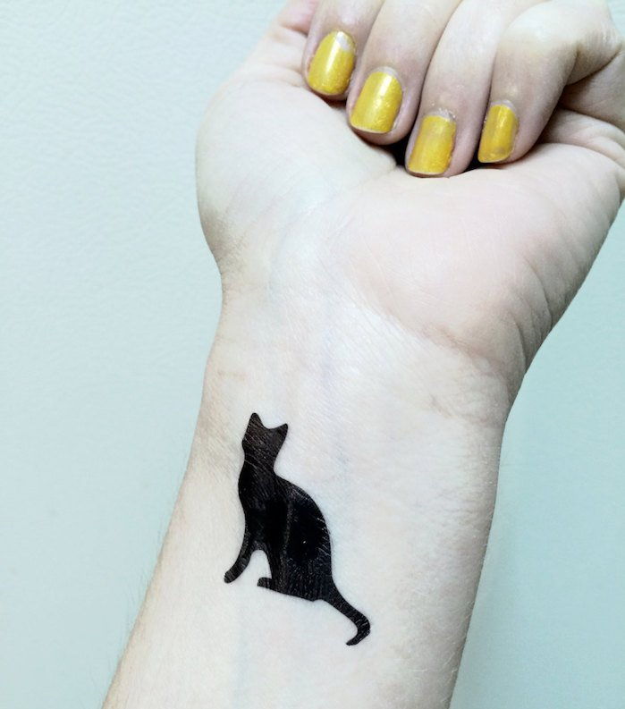 idee buna pentru o pisica tatuaj pe incheietura mainii - aici este o mana, un lac de unghii galben si o pisica neagra