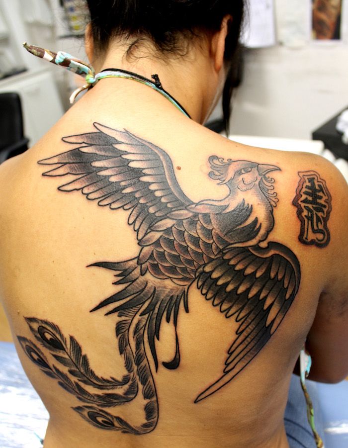 Jauna moteris su juoda tatuiruotė su dideliu juodu plaukiojančiu feniiksu su dviem juodais sparnais su juodos ir baltos spalvos plunksnomis