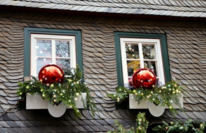 Casă din lemn cu acoperiș înclinat și două ferestre mici cu cadre duble, pervazuri exterioare decorate cu ramuri de conifere cu lumini de zână și două bile imens roșu de pom de Crăciun cu strălucire