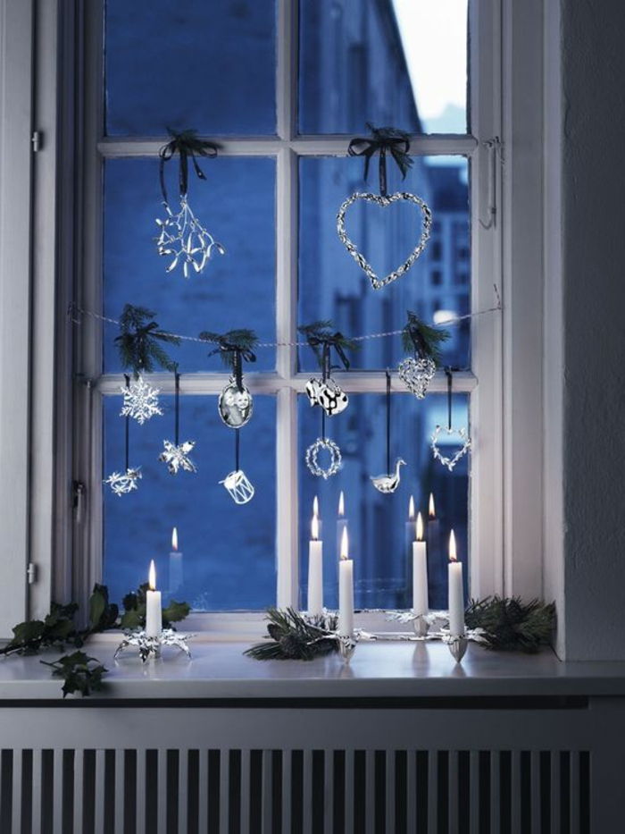 Fensterdeko pentru Ajunul Crăciunului - lumanari albe pe sfesnice elegante, bucăți mici conifer ramuri, fereastra ghirlanda de figurine de sticlă, element decorativ în formă de inimă, cu lumini LED-uri mici și un panou de încălzire panglică verde din lemn