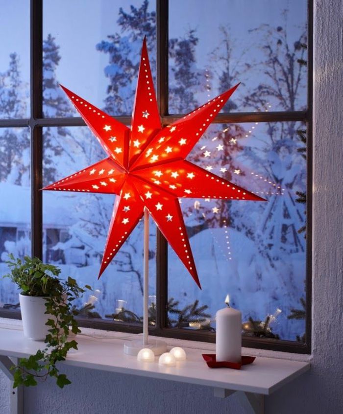 Stojaca lampa s červeným papierovou zástenu v tvare hviezdy, osvetlený sviečka z bieleho vosku, zelená liana v bielom kvetináči z keramiky, zimná krajina s množstvom snehu