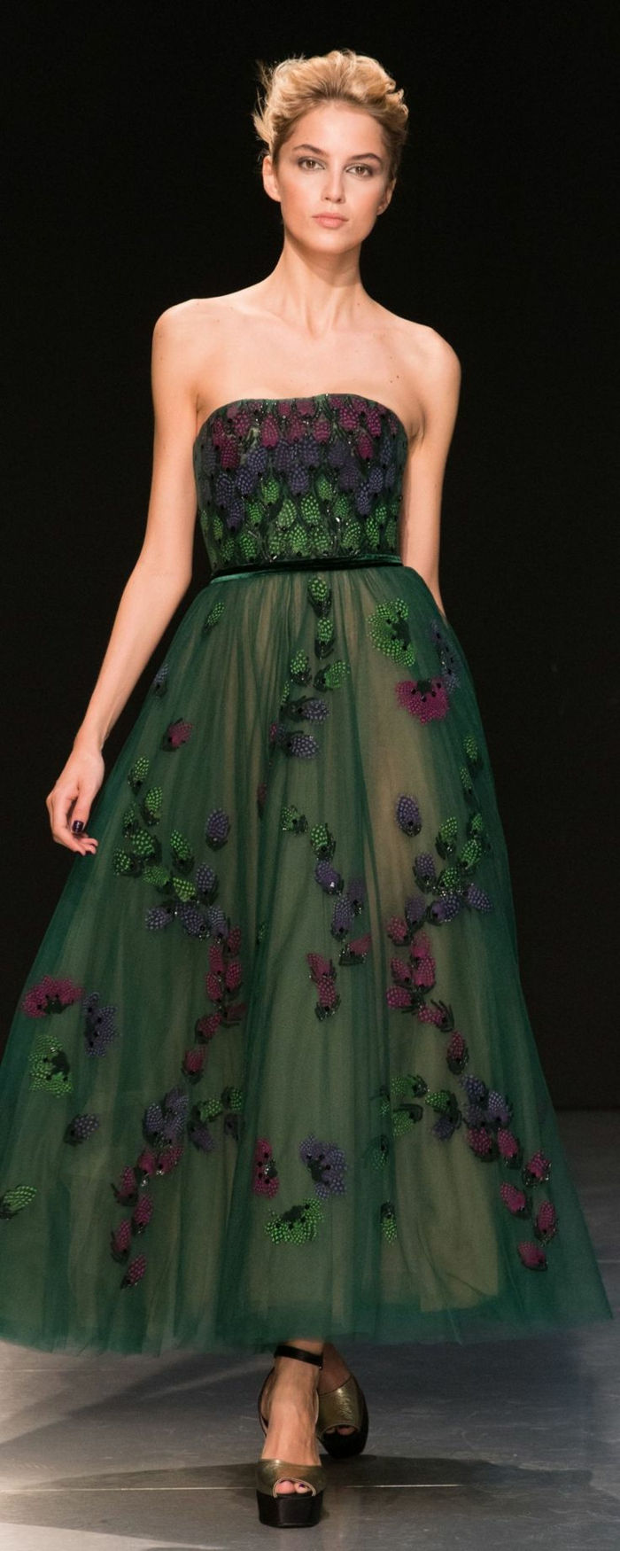 lång mörkgrön stropplös klänning med smalt svart bälte och massor av blå, lila och gröna applikationer, i kombination med plattformsandaler i guldfärg