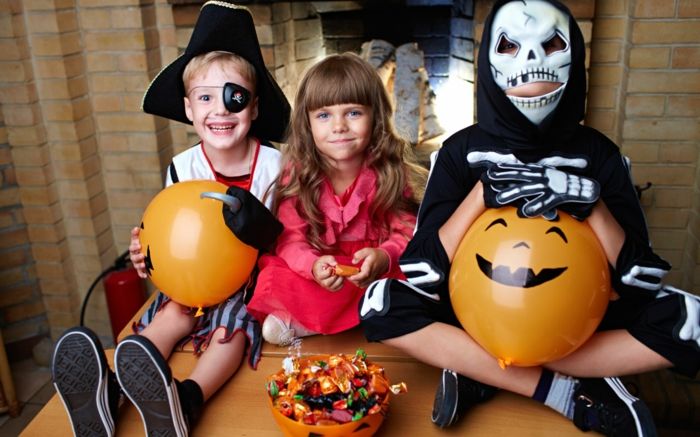 Trys mieli vaikai su Helovino kostiumais, oranžiniai balionai kaip moliūgai, puodelis, pilnas saldumynų