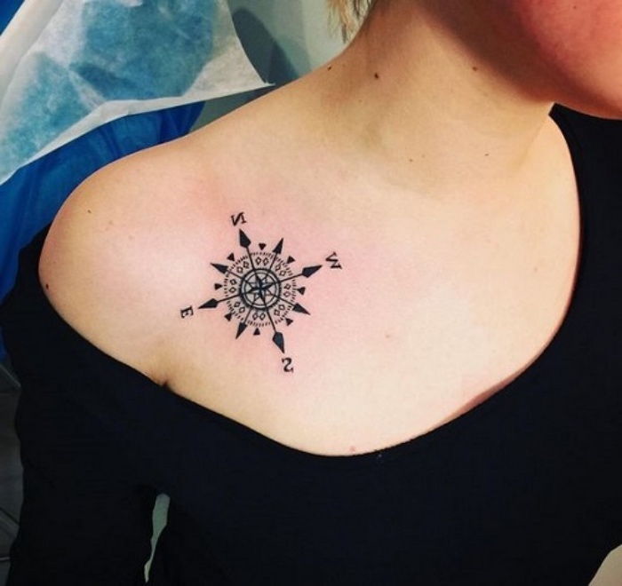 Jest to mały czarny tatuaż z małym czarnym kompasem na ramieniu kobiety