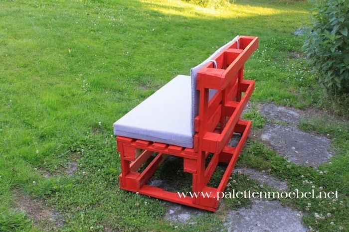 malá červená záhradná lavica vyrobená z starých europalet s purpurovými vankúšmi - záhrada s trávou, zelenými rastlinami a šedými kameňmi