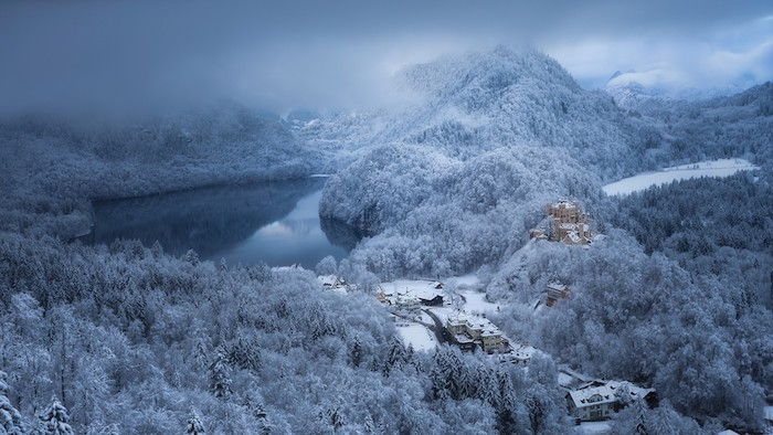 imagini frumoase de iarnă - o pădure cu mulți copaci cu zăpadă și lac și un mic castel galben - cer cu multe nori gri