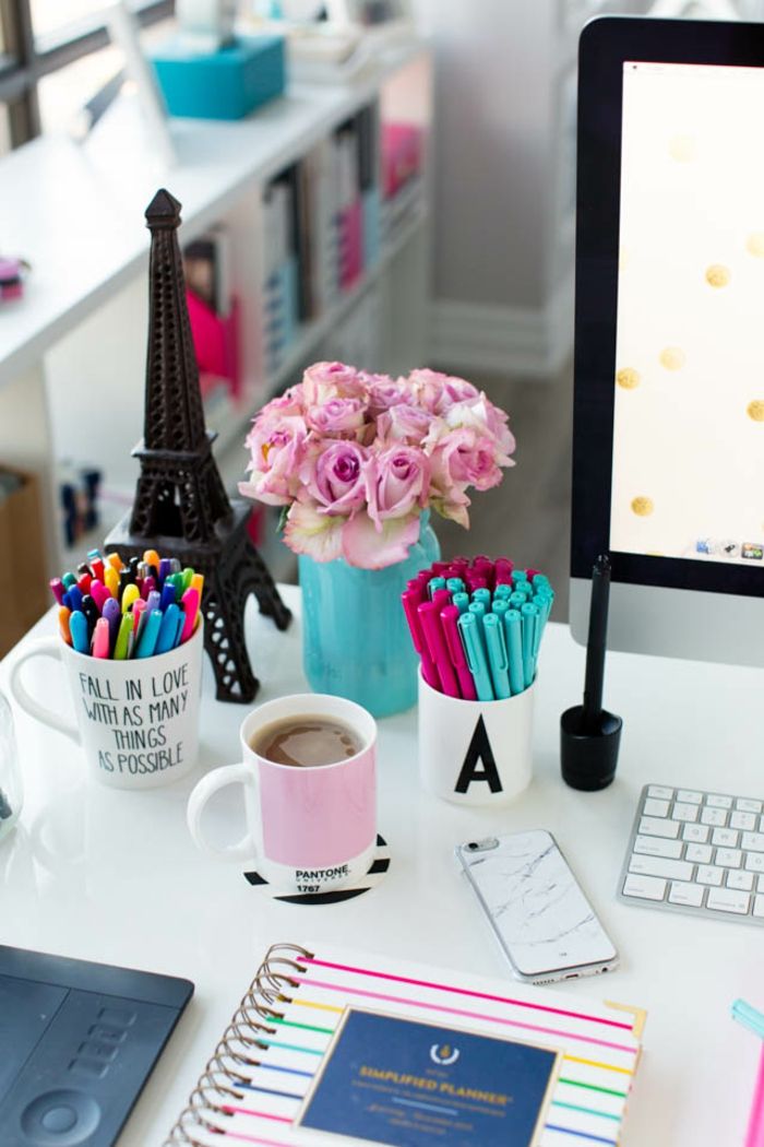 birou alb, buchet de trandafiri în borcanul zidar albastru, cești ca suport pentru stilou, dosar colorat, ceașcă de ceai, PC și telefon mobil