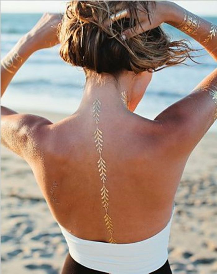 tatuaj idei subtile în culoarea aurie pe spatele femeii cu păr blond pe plajă