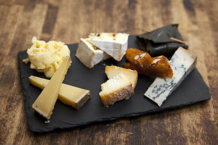 čierna lávová kamenná servírovacia misa položená na drevenom povrchu, dva druhy tvrdého krupicového syra, kúsok modrého syra s trojuholníkovým tvarom, viničový listový rolád s párátkom