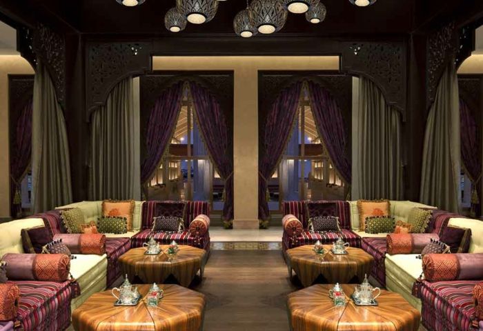 Cafe din Maroc cu patru mese din lemn, cu formă rotundă, Eckcouchen confortabil, cu țesături colorate, pătrate mici și perne rotunde, perdele grele care se încadrează în, iluminat de designer rotund gri și violet, plafon cu sculpturi