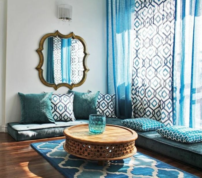 typowy marokański wypoczynkową w odcieniach niebieskim i białym, miękkim wzór dywanu w trzech kolorach, duże lustro na ścianie z nieregularnym kształcie i pozłacanej ramie z przestarzałych wygląd, małe ścianki lampy o niskim okrągłym stoliku z drewna, niebieskim wazonie szkła z okrągłym kształtem, trzy długie półprzezroczyste zasłony niebieski