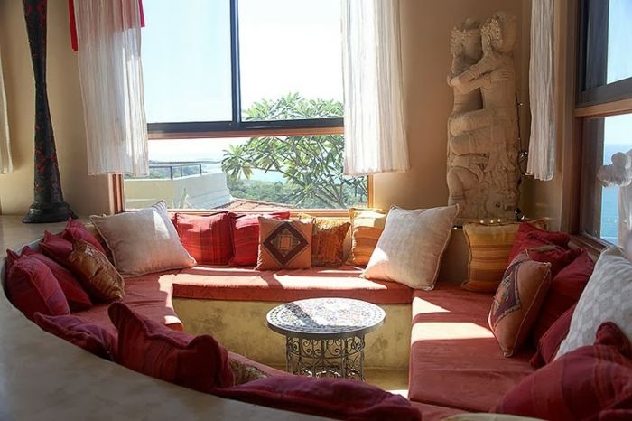 Kaffe hjørne i stuen, terracotta sofa med oval form, rund metall bord med mange ornamenter, to store vinduer med korte hvite gardiner, stein statue i høyre hjørne av rommet
