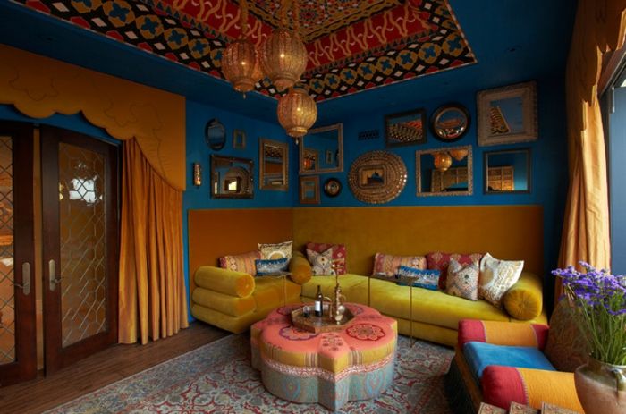blå vegger med masse speil og bilder, gul polstret sofaen i venstre hjørne, wallcovering møbeltrekk, som fungerer som en rygg, oval pute bord i form av en blomst, stoppede stol i mange farger, store mønstre teppebelagte i blått og terrakotta, glassdør med to vinger, dør med oransje gardin tak med dekorative nisje, lavendel bukett i en keramisk fartøy