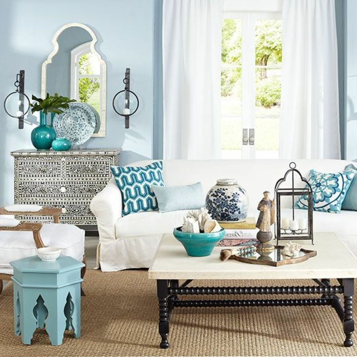 Svetainė su šviesiai mėlynos sienos, ilgi baltos užuolaidos, dekoratyvinis veidrodis su siauru baltu rėmeliu, dekoratyvinės lėkštės dviejų spalvų, žurnalinis staliukas pagamintas iš medžio, dažytos šviesiai mėlyna, balta stalo su juoda medžio kojų ahagoniholz, modelis pagalvės mėlynai, daug dekoratyvinių elementų ant stalo