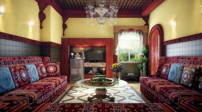 Marokkansk stue med flatskjerm-TV i nisje, gule vegger med mønstret tapet i blått, indirekte veggbelysning i stuen, krystallkroner, rund glassbord med lave treben, hjørnesofa med marokkanske stoffer
