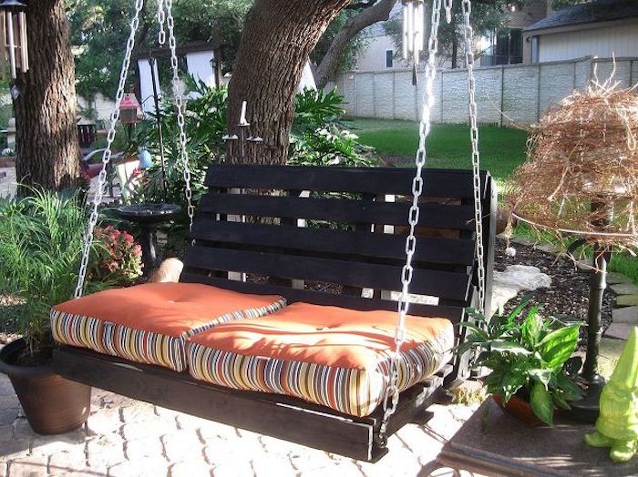 Hojdací stojan z reťazí s oranžovými vankúšmi a hnedou lavicou v záhrade