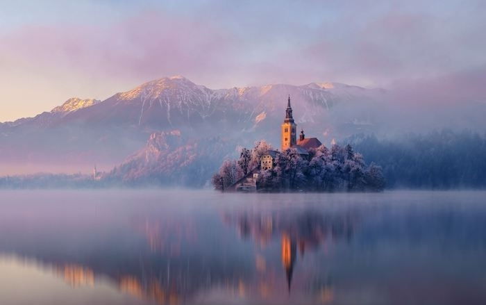 romantiška žiemos scena - ežeras su sala su bažnyčia, nedideli namai ir medžiai - kalnai su sniego ir rožinės spalvos debesimis