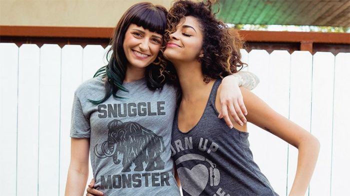Hjemmelaget salg - to unge kvinner med T-skjorter med trykk som klemmer hverandre foran et hvitt gjerde