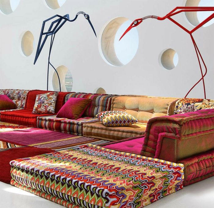 Kanapa z kolorowych tkanin o szalonych wzorach, poduszki w stylu kline, pluszowe dywany w jasnych kolorach, ściana z okrągłymi wnękami, dwie dekoracyjne figury dźwigów