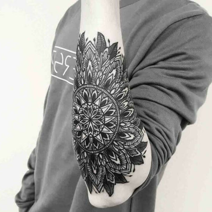 Uomo con un complicato tatuaggio all'avambraccio con un grande mandala in colore nero con molte foglie e cerchi, camicetta grigia a maniche lunghe con stampa di lettere bianche