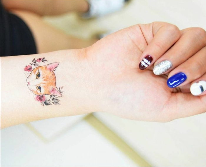 O mână, un deget, un lac de unghii colorat, un tatuaj de pisică pe încheietura mâinii, o pisică mică, flori roșii