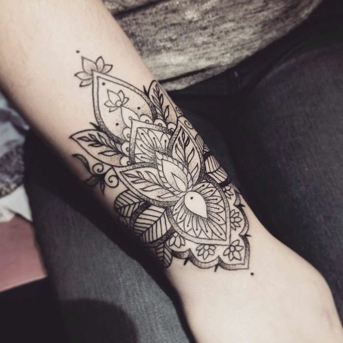 Tatuaż na nadgarstku z dwoma małymi szarymi kwiatami lotosu, spirale i wieloma liśćmi, ornamentem w małe kropki, czarnymi dżinsami i szarą bluzką