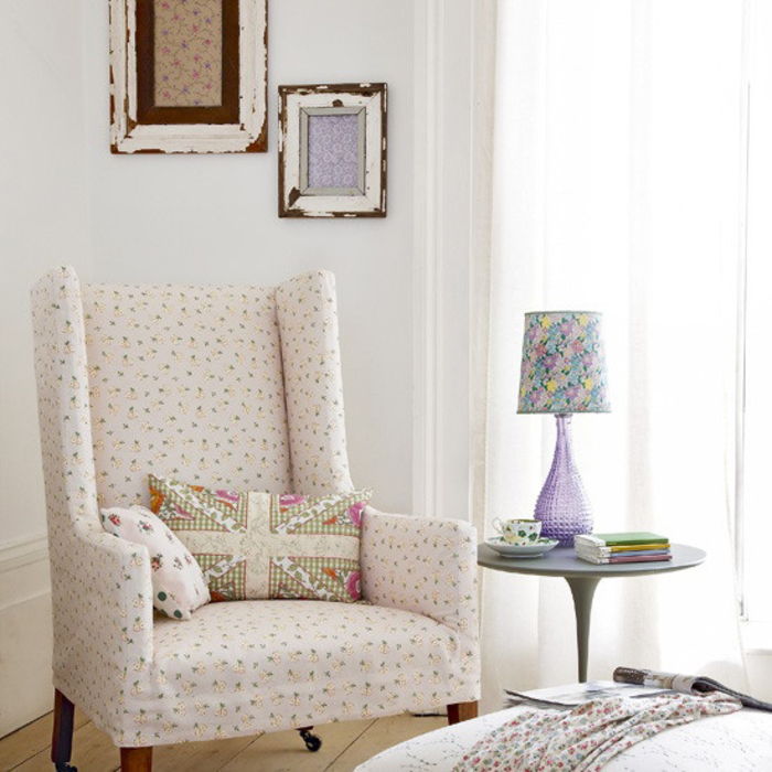 Stilingas gyvenamasis kambarys, balta kėdė ir dekoratyvinė pagalvė, violetinė naktinė lempa, puodelis, gėlių raštas