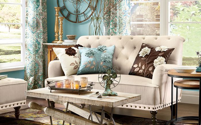 Stilingas gyvenamasis kambarys, sukuriantis romantišką išvaizdą, dekoratyvinė pagalvė su gėlių raštu, medinis stalas, žvakių laikiklis, retro ventiliatorius