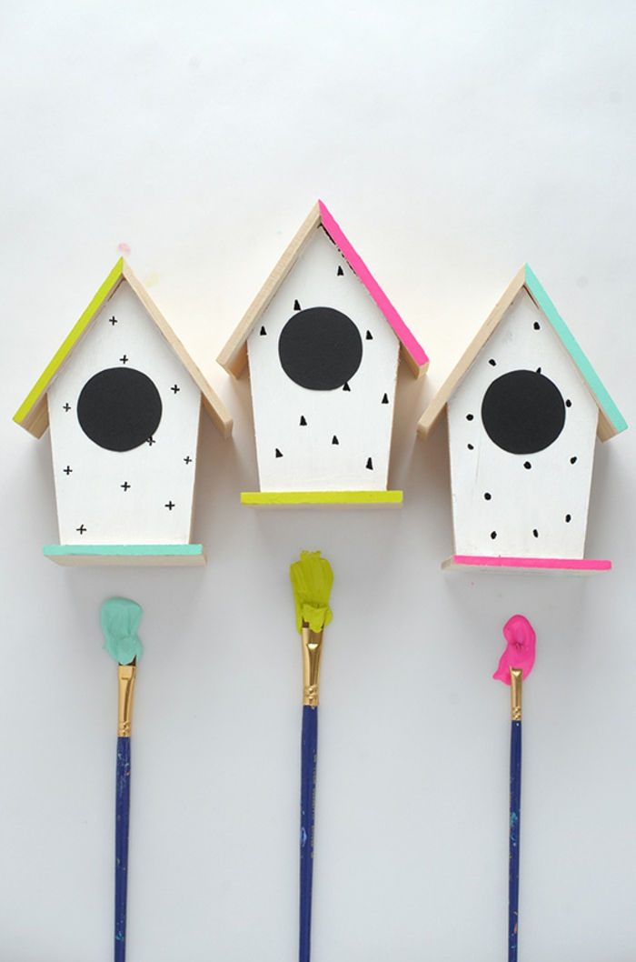 três caixas alimentadoras de pássaros feitas de madeira, paredes brancas decoradas com pontos, cruzes e triângulos, telhados coloridos